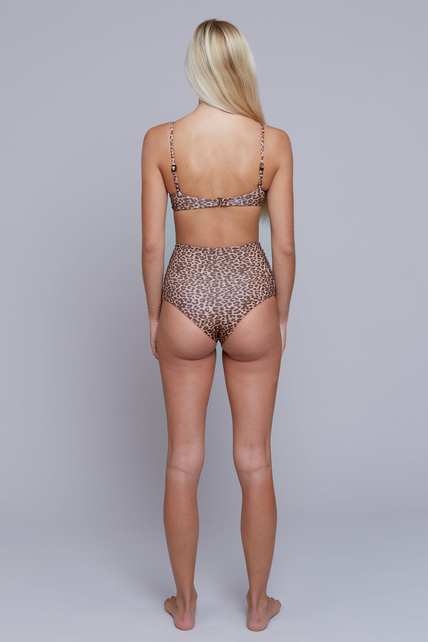 A woman stands with her back to the camera wearing a leopard-print bikini set against a plain light gray studio background.