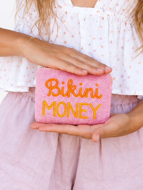 A person holding a pink beaded clutch bag with orange embroidered text that reads 'Bikini MONEY'.