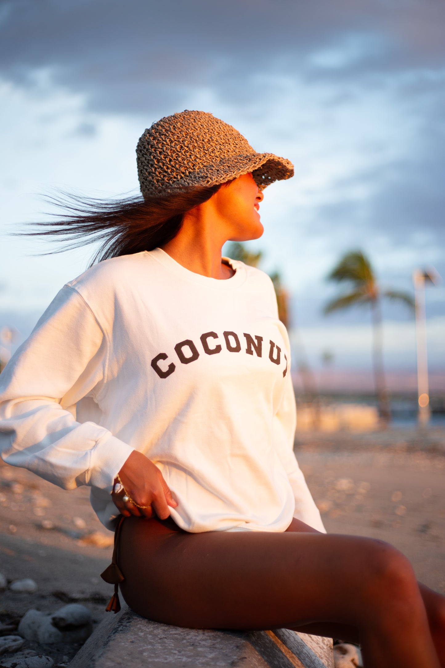 A woman sitting on a beach wearing a white sweatshirt with 'COCONUT' printed in brown across the chest, a woven straw hat, and dark brown shorts, captured in warm golden hour light.