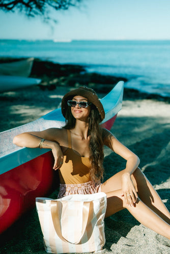 Woman relaxing beside a canoe on a tropical beach with a Coconut Ave tote bag, wearing lightweight resort wear and sun hat for an island getaway.