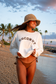 A woman walking on a beach at sunset wearing a cream sweatshirt with 'COCONUT' printed across the chest, a woven straw hat, and a brown bikini. Palm trees and the ocean are visible in the background.