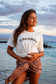 A woman standing on a beach at sunset wearing a white graphic t-shirt with 'COCONUT' printed in brown across the chest, paired with a metallic bronze wrap skirt.