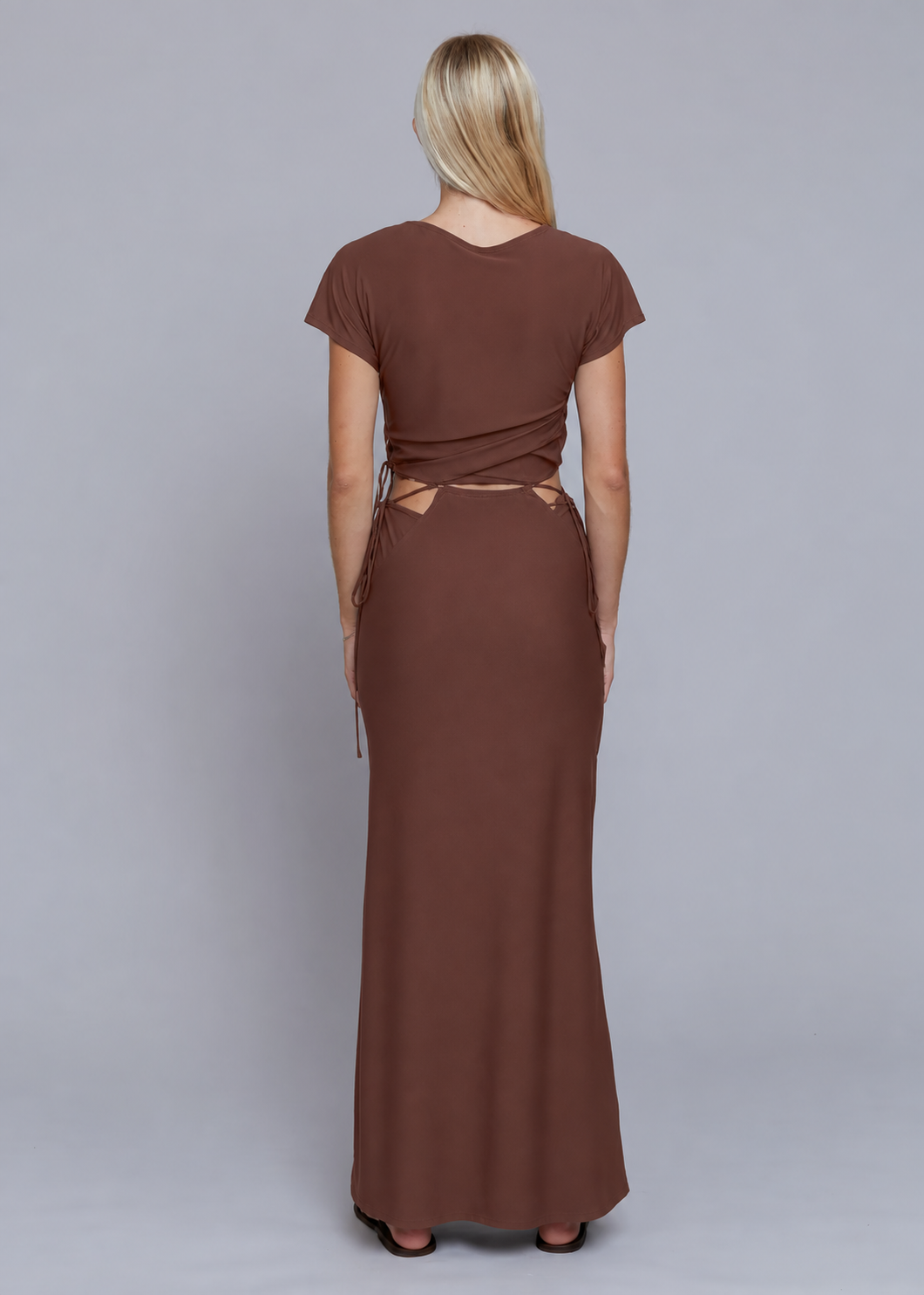 A woman viewed from the back wearing a brown short-sleeve maxi dress with a wrap-style back and tie details, standing against a plain gray studio background.