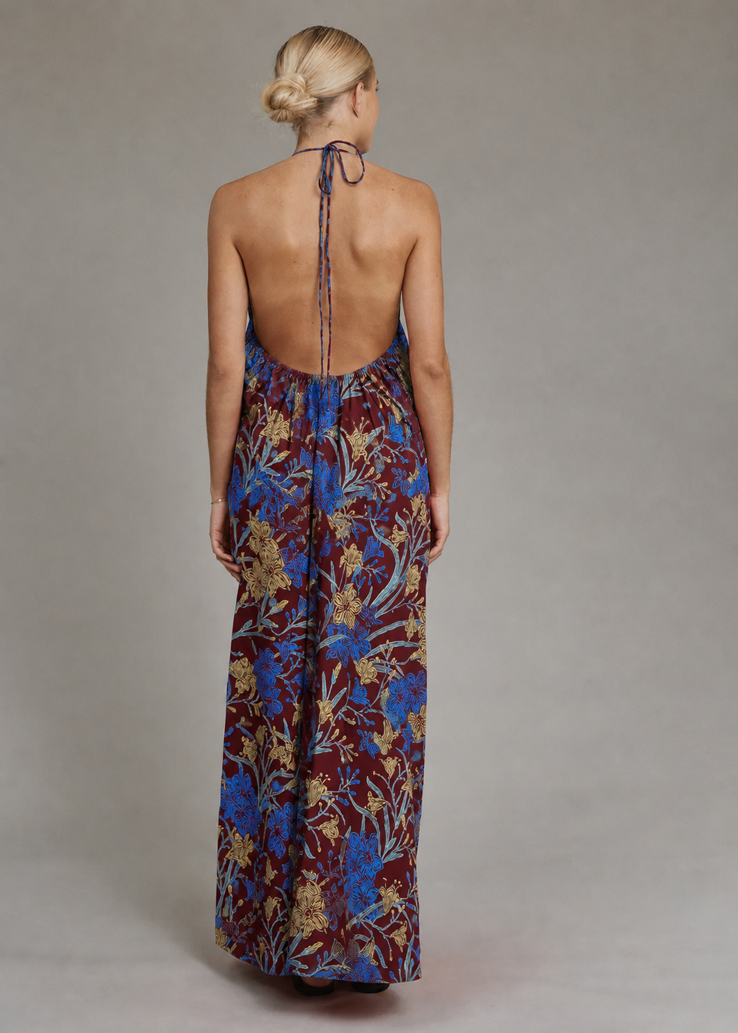 A woman viewed from the back wearing a sleeveless, open-back maxi dress with a blue and gold floral pattern on a dark burgundy base, tied at the neck, set against a plain gray studio background.