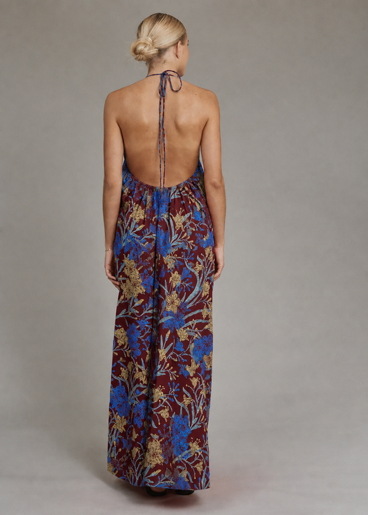 A woman viewed from the back wearing a sleeveless, open-back maxi dress with a blue and gold floral pattern on a dark burgundy base, tied at the neck, set against a plain gray studio background.