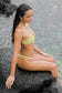 Side view of a woman sitting on volcanic rocks wearing the Coconut Ave Hula Bandeau Top in the Moray print with matching bikini bottoms, styled for beachwear and tropical vacations.