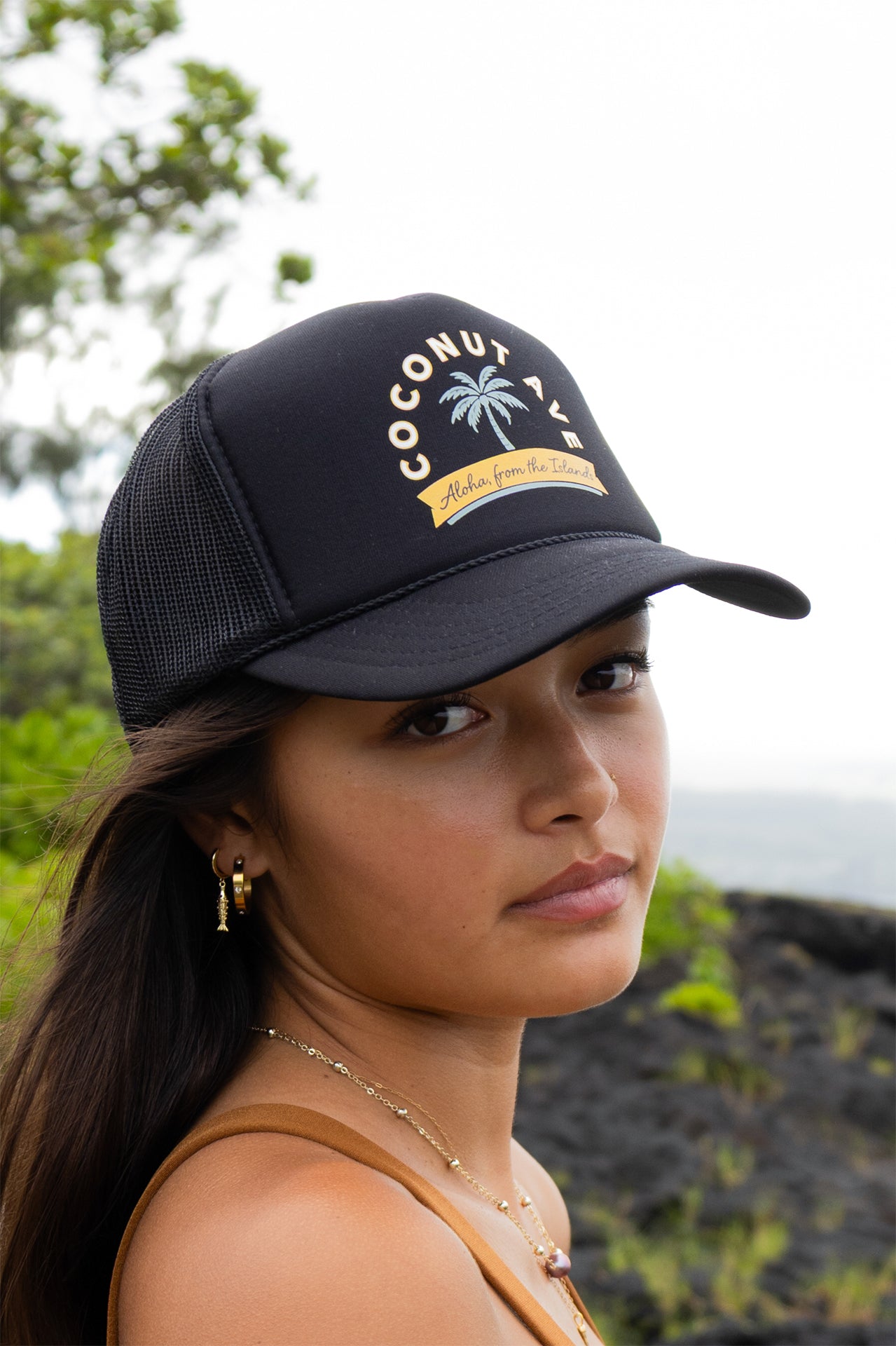 Woman wearing the Coconut Ave Trucker Hat – Aloha From The Islands featuring a black mesh back, structured front panel with embroidered graphic, rope detail, and adjustable snapback closure.