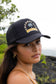 Woman wearing the Coconut Ave Trucker Hat – Aloha From The Islands featuring a black mesh back, structured front panel with embroidered graphic, rope detail, and adjustable snapback closure.