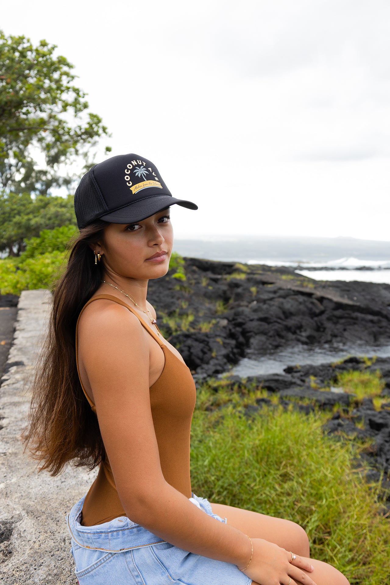 Woman wearing the Coconut Ave Trucker Hat – Aloha From The Islands in an outdoor setting, showcasing the embroidered front graphic, rope accent, mesh back, and adjustable trucker hat fit.