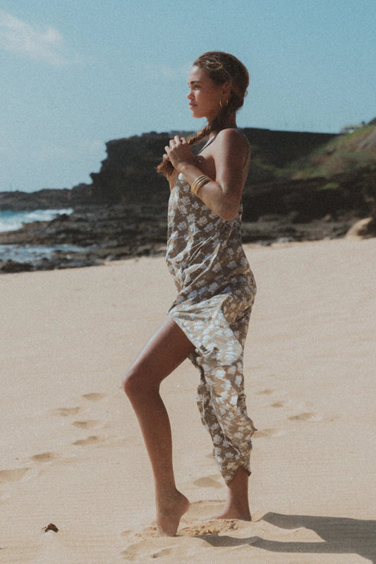 A woman stands barefoot on a sandy beach wearing a sleeveless, patterned wrap-style dress or cover-up in beige, white, and brown tones. The background shows rocks, ocean, and blue sky.