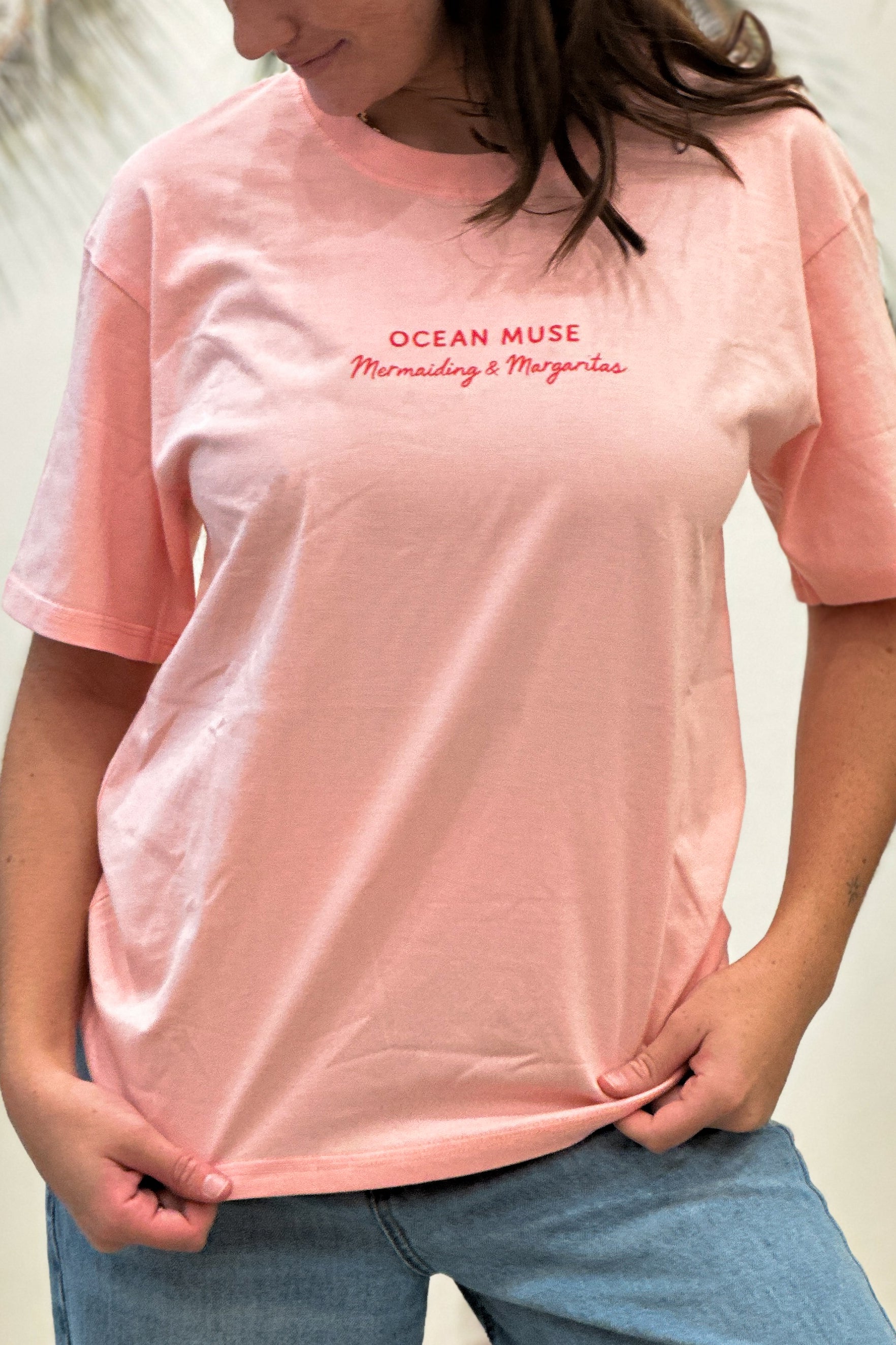 Person wearing a pink t-shirt with 'Ocean Muse' text, standing against a neutral background