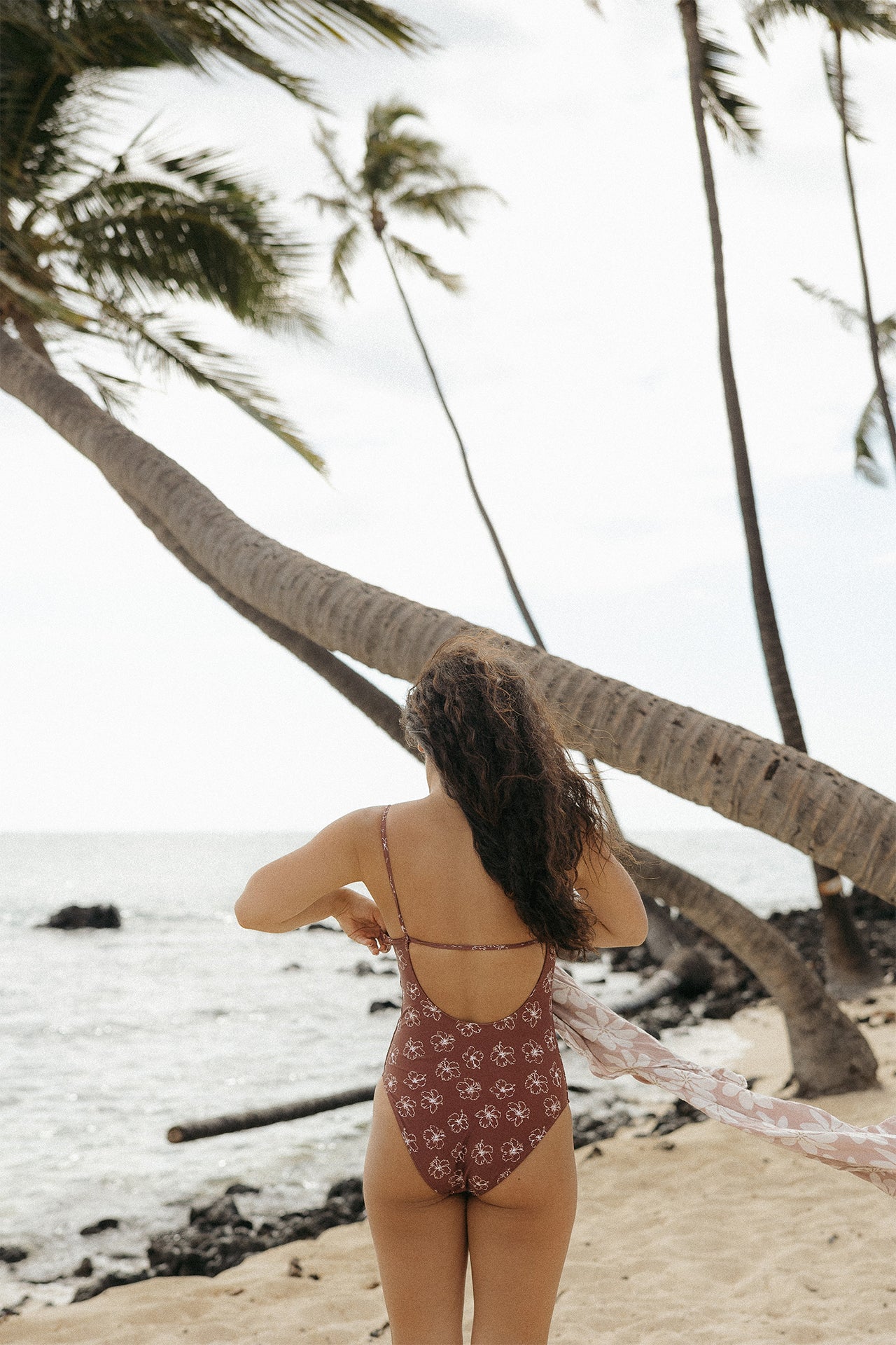 Side view of a woman wearing the Stevie One Piece in Chocolate Bloom in a tropical beach setting