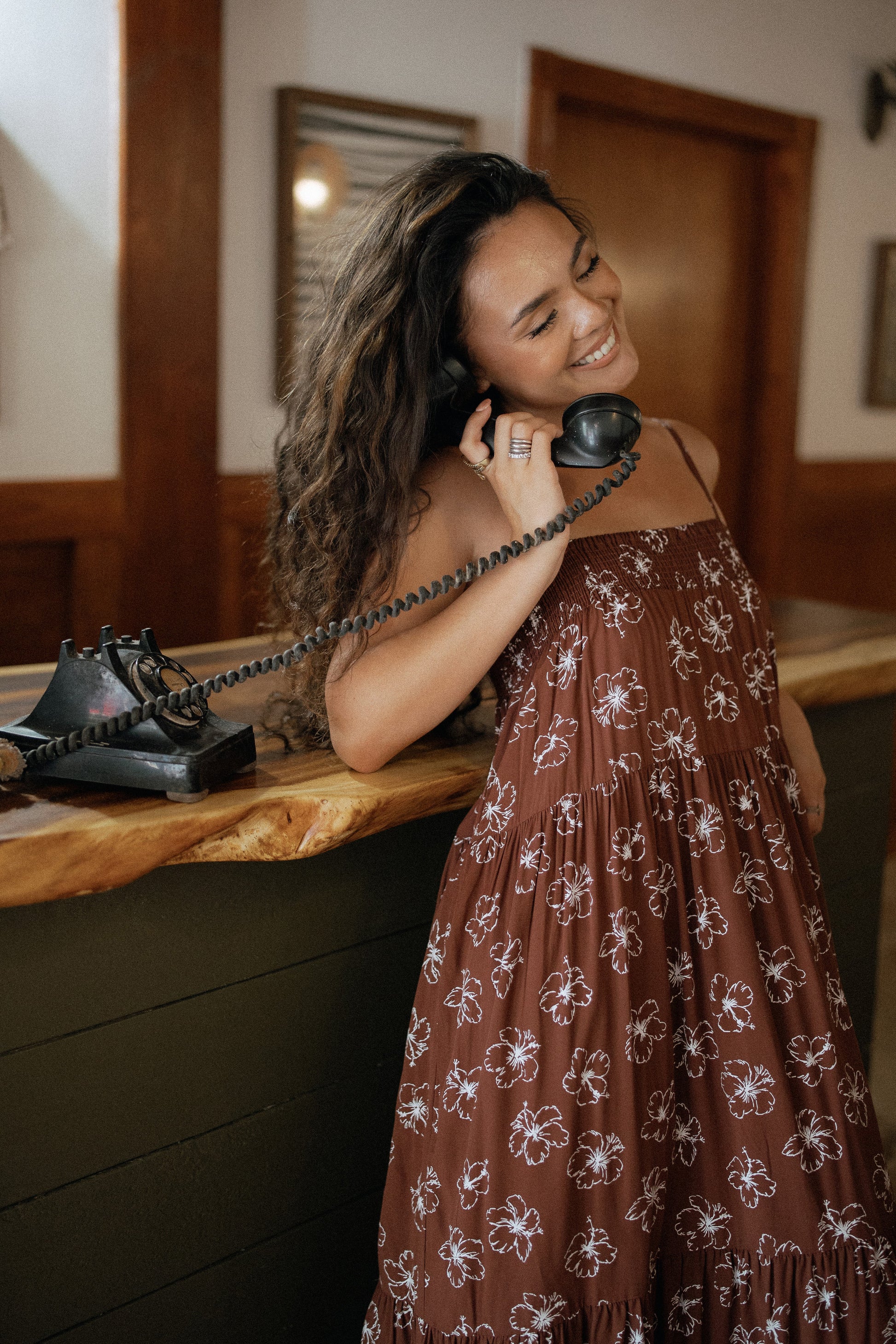 Chocolate Bloom Waikiki Maxi Dress styled with sandals for a relaxed resort dinner in a warm indoor setting.