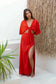 Woman wearing the Coconut Ave Minx Maxi Skirt in Hibiscus with a matching red crop top while standing beside a textured tropical wall. The flowing linen blend maxi skirt features a high side slit, elasticized waistband, and elegant resort wear styling for beach vacations and island getaways.