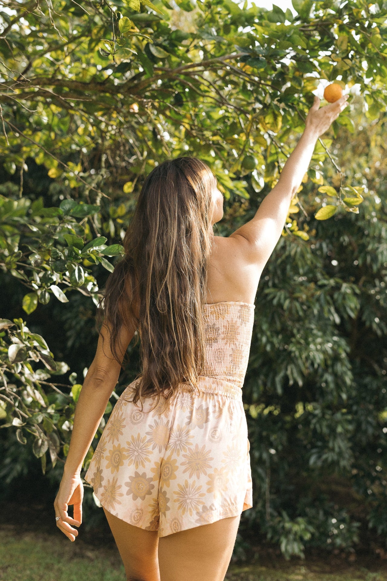Back view of a woman wearing the Coconut Ave Smocked Tube Top and matching Playa Shorts in the Lilikoi Blooms print, showing the lightweight rayon fabric, floral pattern, and coordinated tropical outfit in a garden setting.