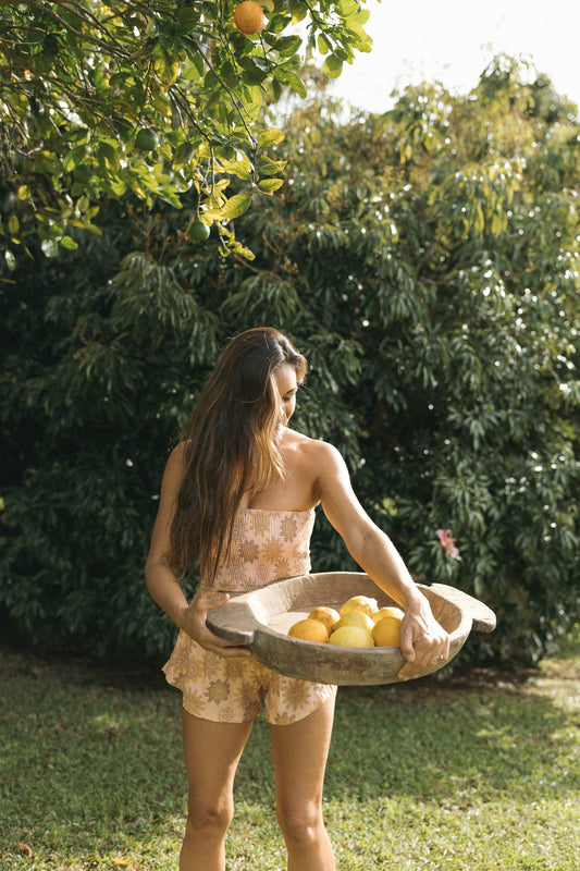 Rear view of a woman wearing the Coconut Ave Smocked Tube Top in Lilikoi Blooms with matching Playa Shorts, showcasing the strapless smocked design, cropped silhouette, and exclusive floral print in an outdoor tropical setting.