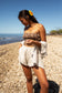 Woman wearing the Coconut Ave Smocked Tube Top in the Sexy Pink print with white linen shorts while standing on a rocky beach. The strapless smocked rayon crop top features Coconut Ave's exclusive tropical print and is styled as a lightweight women's resort wear outfit for beach vacations and summer travel.