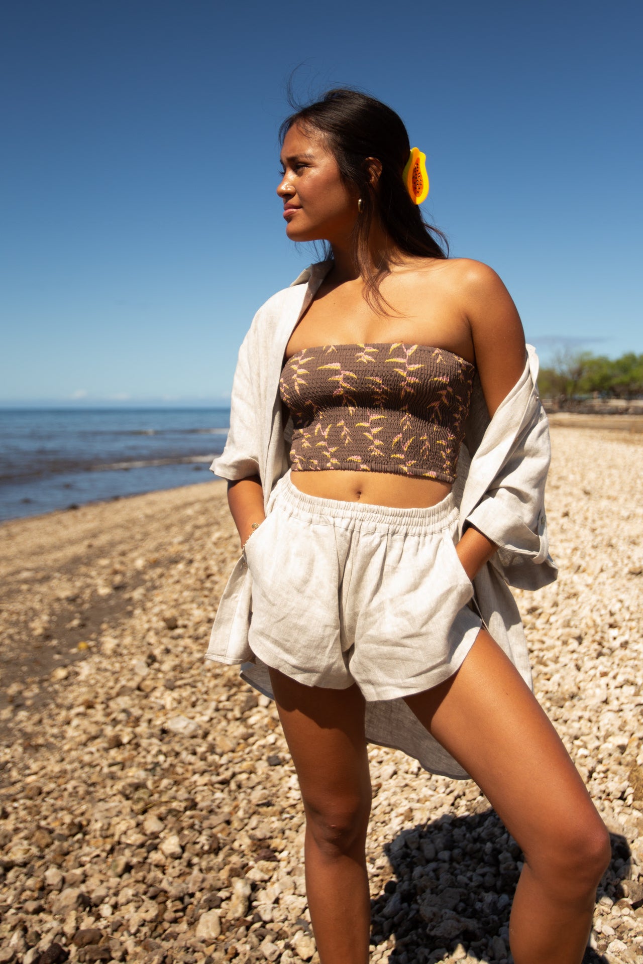 Woman wearing the Coconut Ave Smocked Tube Top in the Sexy Pink print paired with white linen shorts while standing on a rocky shoreline. The image highlights the strapless cropped design, comfortable stretch smocking, and tropical beach styling for women's resort wear.